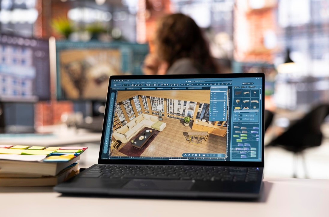 Portátil mostrando un diseño de interiores en 3D con un salón moderno, decorado con un sofá, mesa de café y plantas, en un ambiente de oficina creativa.